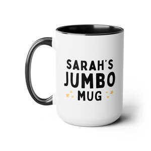 Może przedstawiać: Biały kubek ceramiczny z czarną obw&oacute;dką i uchwytem. Kubek ma czarny napis "SARAH'S JUMBO MUG" z małymi złotymi gwiazdkami po obu stronach napisu.