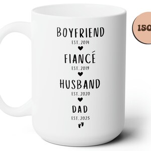 K&ouml;nnte beinhalten: Wei&szlig;e Keramiktasse mit schwarzem Text, der "Boyfriend Est. 2014", "Fianc&eacute; Est. 2019", "Husband Est. 2020" und "Dad Est. 2023" lautet. Die Tasse ist 15 oz.