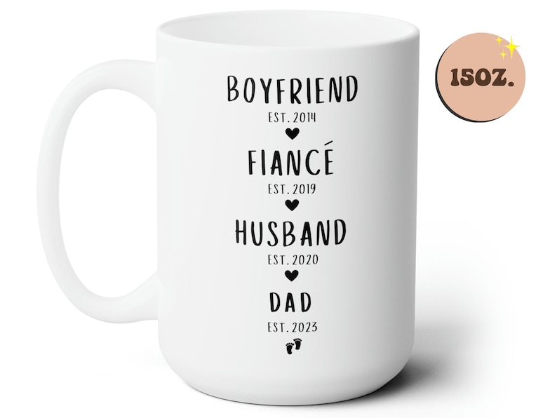 K&ouml;nnte beinhalten: Wei&szlig;e Keramiktasse mit schwarzem Text, der "Boyfriend Est. 2014", "Fianc&eacute; Est. 2019", "Husband Est. 2020" und "Dad Est. 2023" lautet. Die Tasse ist 15 oz.