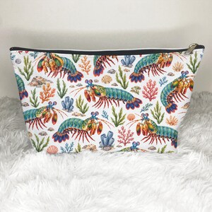 May include: A white zipped pouch featuring a repeating pattern of colourful mantis shrimp, orange coral, green seaweed, and small seashells. The design displays multiple shrimp in shades of blue, green, and orange against a clean white background.