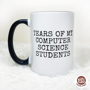 May include: White ceramic mug with a black handle and rim. The mug features the text "TEARS OF MY COMPUTER SCIENCE STUDENTS" in black, bold letters. The mug is sitting on a white, fluffy surface.