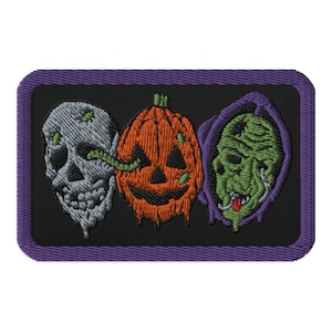 May include: An embroidered patch with a black background and a purple border. The patch features three Halloween-themed faces: a skull, a pumpkin, and a green monster.