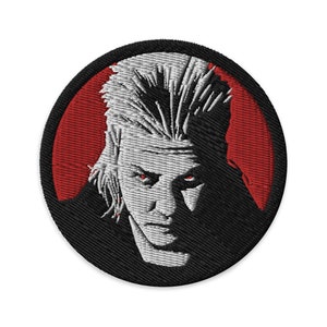 May include: Embroidered patch featuring a black and white portrait of a man with a white, spiky hairstyle. The man has a serious expression and is looking directly at the viewer. The background is a red circle.