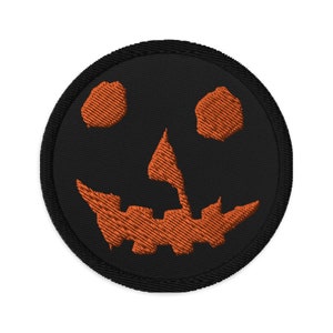 May include: A black embroidered patch with an orange jack-o'-lantern face. The patch is circular and has a black border.