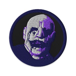 Puede incluir: Parche bordado circular con un retrato estilizado de un rostro. El rostro está representado en hilo blanco y morado sobre un fondo negro, rodeado por un borde azul oscuro. El diseño es circular.