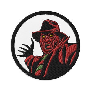 Può includere: Una toppa ricamata rossa e nera con un personaggio di un film horror. Il personaggio indossa una camicia a righe rosse e nere, un cappello nero e ha un'espressione minacciosa. La toppa è circolare e ha un bordo nero.