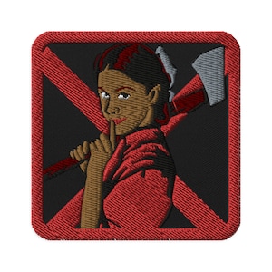 May include: A red embroidered patch with a black background. The patch features a woman with red hair holding an axe. The woman is wearing a red shirt and has a finger to her lips. The patch is surrounded by a red border.