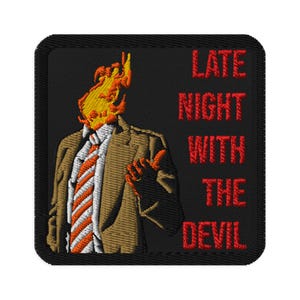 May include: Embroidered patch with a black background and red text that reads "Late Night With The Devil". A cartoon image of a man with a flaming head wearing a brown suit and a striped tie is in the left side of the patch.