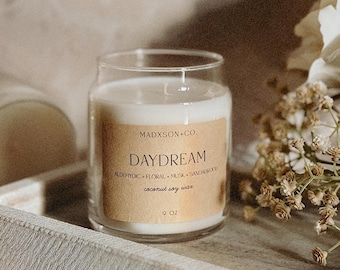 May include: A clear glass jar candle with a beige label that reads "DAYDREAM" and "coconut soy wax." The candle is set on a light gray tray with dried flowers and a white flower in the background. The candle is 9 oz.