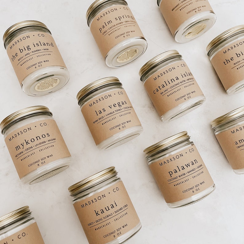 May include: Ten glass candle jars with gold lids and brown paper labels. Each label has the name of a location and a scent description. The candles are arranged in a grid pattern on a white surface. The candles are all 8 oz.