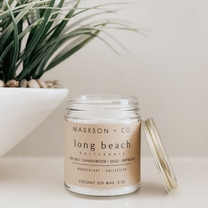 May include: A glass candle jar with a brown paper label that reads "MADXSON + CO. long beach CALIFORNIA SEA SALT SANDALWOOD SAGE DRIFTWOOD WANDERLUST COLLECTION COCONUT SOY WAX 8 OZ". The candle jar has a gold lid.