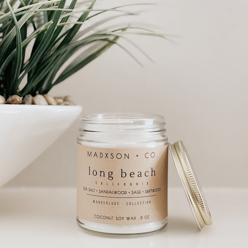 May include: A glass candle jar with a brown paper label that reads "MADXSON + CO. long beach CALIFORNIA SEA SALT SANDALWOOD SAGE DRIFTWOOD WANDERLUST COLLECTION COCONUT SOY WAX 8 OZ". The candle jar has a gold lid.