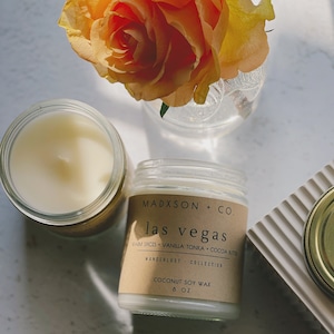 May include: A brown candle jar with a white label that reads "MADXSON + CO. las vegas WARM SPICES + VANILLA TONKA + COCOA BUTTER WANDERLUST COLLECTION COCONUT SOY WAX 8 OZ". The candle is sitting on a white surface with a rose in the background.