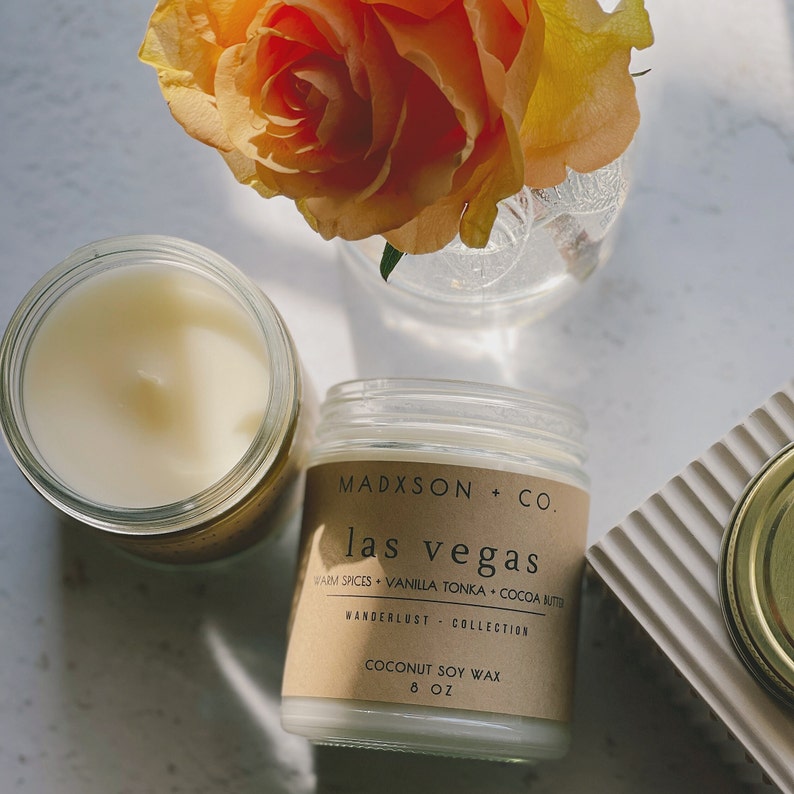 May include: A brown candle jar with a white label that reads "MADXSON + CO. las vegas WARM SPICES + VANILLA TONKA + COCOA BUTTER WANDERLUST COLLECTION COCONUT SOY WAX 8 OZ". The candle is sitting on a white surface with a rose in the background.