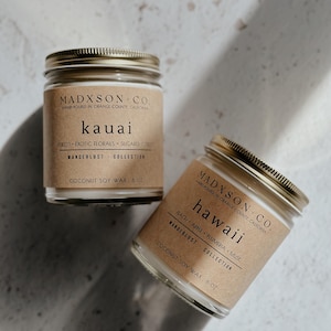 May include: Two glass candle jars with brown paper labels. The labels have gold text that reads "MADXSON-CO. HAND-POURED IN ORANGE COUNTY, CALIFORNIA". The first jar is labeled "kauai" and the second jar is labeled "hawaii". Both jars are filled with white wax and have gold lids.