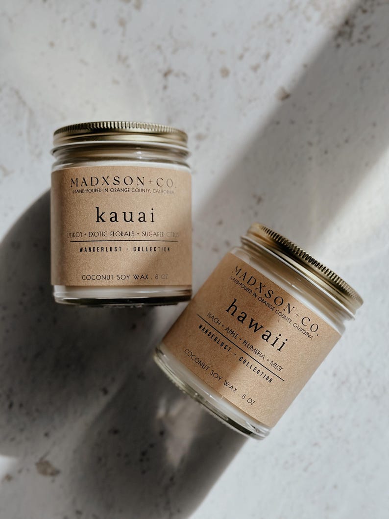 May include: Two glass candle jars with brown paper labels. The labels have gold text that reads "MADXSON-CO. HAND-POURED IN ORANGE COUNTY, CALIFORNIA". The first jar is labeled "kauai" and the second jar is labeled "hawaii". Both jars are filled with white wax and have gold lids.