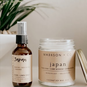 May include: A brown glass bottle with a black spray nozzle and a label that reads "Madxson + Co. Japan Asian Pear Cherry Blossom Sandalwood Room + Linen Spray 2 fl oz 159 ml". Next to the bottle is a clear glass jar with a brown label that reads "Madxson + Co. Japan Asian Pear Cherry Blossom Sandalwood Wanderlust Collection Coconut Soy Wax 8 oz".