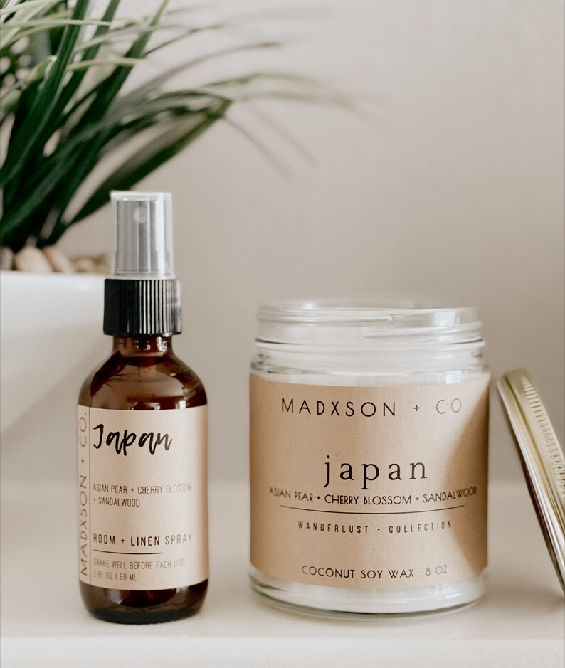 May include: A brown glass bottle with a black spray nozzle and a label that reads "Madxson + Co. Japan Asian Pear Cherry Blossom Sandalwood Room + Linen Spray 2 fl oz 159 ml". Next to the bottle is a clear glass jar with a brown label that reads "Madxson + Co. Japan Asian Pear Cherry Blossom Sandalwood Wanderlust Collection Coconut Soy Wax 8 oz".