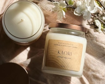May include: Two glass jar candles with white wax and copper-colored lids. One candle has a label that reads "KAORI coconut soy wax" and "MADXSON + CO." The candles are on a beige cloth with white flowers.