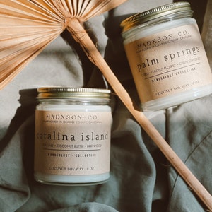 May include: Two glass candle jars with brown paper labels. The labels have the text "MADXSON CO. HAND-POURED IN ORANGE COUNTY, CALIFORNIA" and the names of the scents: "catalina island" and "palm springs". The candles are on a green fabric surface.