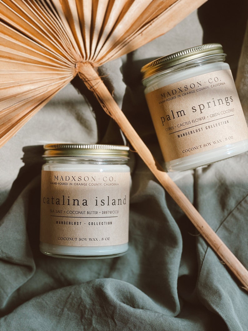 May include: Two glass candle jars with brown paper labels. The labels have the text "MADXSON CO. HAND-POURED IN ORANGE COUNTY, CALIFORNIA" and the names of the scents: "catalina island" and "palm springs". The candles are on a green fabric surface.
