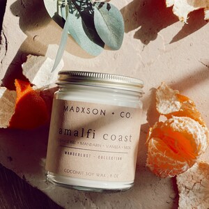 May include: A glass candle jar with a brown label that reads "MADXSON + CO. amalfi coast ORANGE PEEL MANDARIN VANILLA MUSK WANDERLUST COLLECTION COCONUT SOY WAX, 8 OZ". The candle is white and the jar has a gold lid. The candle is surrounded by orange peels.