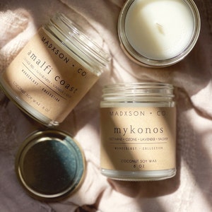 May include: Two glass candle jars with brown paper labels. The labels have the text "MADXSON + CO." and the names of the scents, "amalfi coast" and "mykonos". The candles are on a light pink fabric background.
