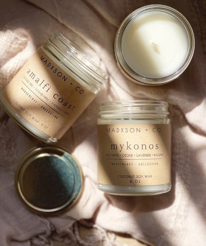May include: Two glass candle jars with brown paper labels. The labels have the text "MADXSON + CO." and the names of the scents, "amalfi coast" and "mykonos". The candles are on a light pink fabric background.