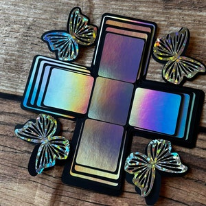 以下が含まれることがあります： イリデセントパネルと4つの装飾的な蝶の切り抜きが付いた、開いた黒いエクスプロージョンボックス。ボックスはカラフルなインテリアを明らかにします。蝶は、複雑な切り抜きデザインのシルバーです。