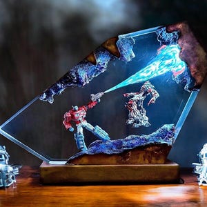 May include: A clear, angled resin sculpture featuring a red and blue robot figure shooting a blue energy beam at another robot. The sculpture is set on a wooden base, with two silver robot figures on either side. The background is dark.