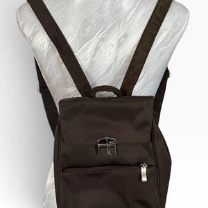 May include: A dark brown nylon backpack featuring a top flap with a silver-toned metal push-lock closure, a small top handle, and adjustable shoulder straps. The front includes a horizontal zippered pocket with a silver-toned metal pull tab.