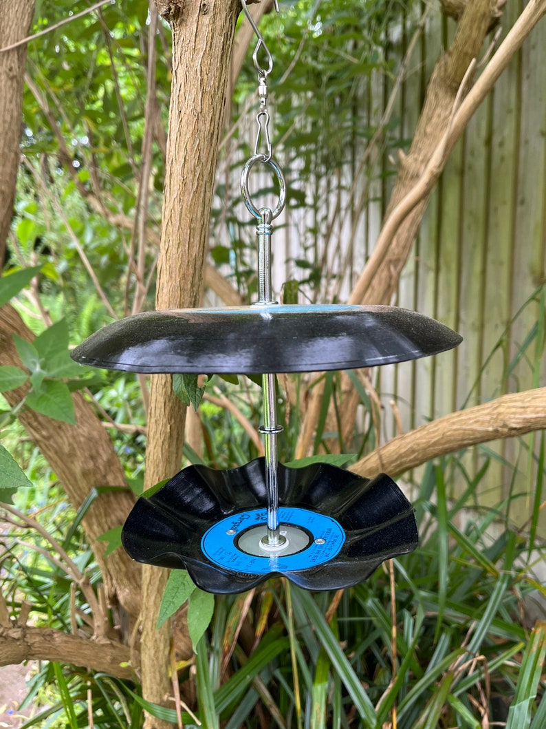 Puede incluir: Un comedero para p&aacute;jaros hecho a mano con dos discos de vinilo negros reciclados. El comedero tiene una varilla de metal plateado y un mecanismo para colgarlo. Los discos est&aacute;n unidos a la varilla, creando un dise&ntilde;o de dos niveles. El comedero para p&aacute;jaros est&aacute; colgado de una rama.
