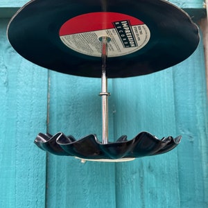 Puede incluir: Un comedero para p&aacute;jaros hecho a mano con un disco de vinilo reciclado. El disco negro tiene una etiqueta roja con texto blanco y un borde festoneado. Cuelga de una varilla y un gancho de metal plateado.