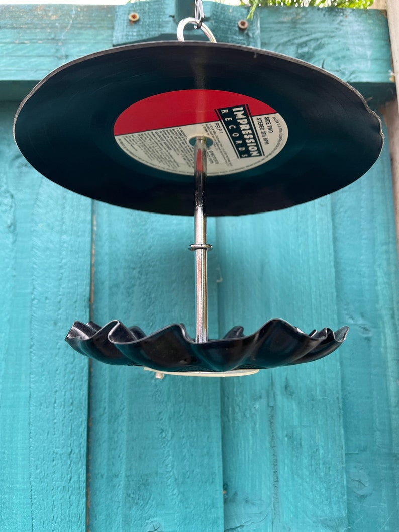 Puede incluir: Un comedero para p&aacute;jaros hecho a mano con un disco de vinilo reciclado. El disco negro tiene una etiqueta roja con texto blanco y un borde festoneado. Cuelga de una varilla y un gancho de metal plateado.