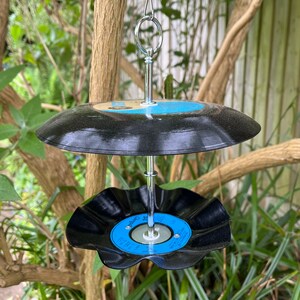 Puede incluir: Un comedero para p&aacute;jaros de dos niveles hecho de discos de vinilo reciclados. Los discos negros tienen etiquetas azules y blancas y est&aacute;n conectados por una varilla de metal plateado y un lazo para colgar. El comedero est&aacute; suspendido de una rama de &aacute;rbol.