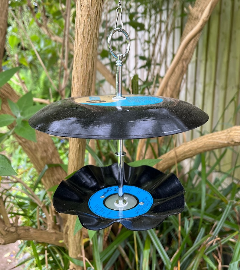 Puede incluir: Un comedero para p&aacute;jaros de dos niveles hecho de discos de vinilo reciclados. Los discos negros tienen etiquetas azules y blancas y est&aacute;n conectados por una varilla de metal plateado y un lazo para colgar. El comedero est&aacute; suspendido de una rama de &aacute;rbol.