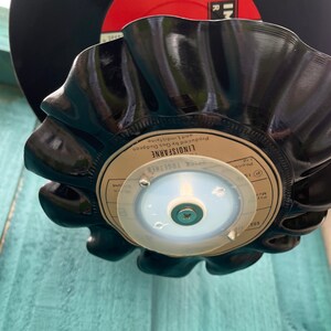 Puede incluir: Un cuenco negro de vinilo reciclado con un borde ondulado. El cuenco est&aacute; hecho de un disco reutilizado, con la etiqueta original visible. El cuenco est&aacute; montado sobre una base de pl&aacute;stico transparente con un tornillo. Un segundo disco es visible en el fondo.
