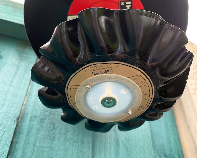 Puede incluir: Un cuenco negro de vinilo reciclado con un borde ondulado. El cuenco est&aacute; hecho de un disco reutilizado, con la etiqueta original visible. El cuenco est&aacute; montado sobre una base de pl&aacute;stico transparente con un tornillo. Un segundo disco es visible en el fondo.