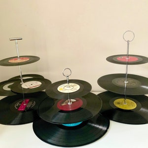 May include: Three-tiered serving stands made from vintage vinyl records. The stands have a silver metal rod and circular handle. Each tier holds black records with colourful labels, creating a unique display for desserts or snacks.