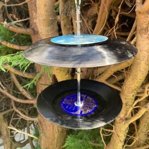 Puede incluir: Un comedero para p&aacute;jaros de dos niveles hecho de discos de vinilo negros reciclados. Los discos est&aacute;n suspendidos por una varilla de metal plateado, con una etiqueta azul y blanca visible en el disco inferior. Cuelga de una rama.