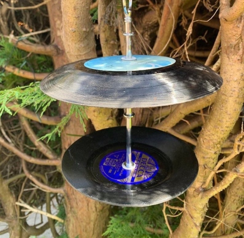 Puede incluir: Un comedero para p&aacute;jaros de dos niveles hecho de discos de vinilo negros reciclados. Los discos est&aacute;n suspendidos por una varilla de metal plateado, con una etiqueta azul y blanca visible en el disco inferior. Cuelga de una rama.