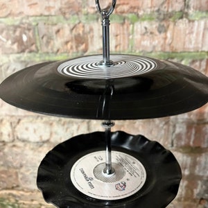 Puede incluir: Un comedero para p&aacute;jaros de dos niveles hecho de discos de vinilo reciclados. El nivel superior presenta un disco negro con un dise&ntilde;o en espiral blanco, mientras que el nivel inferior tiene una etiqueta con texto. Suspendido por una cadena plateada.