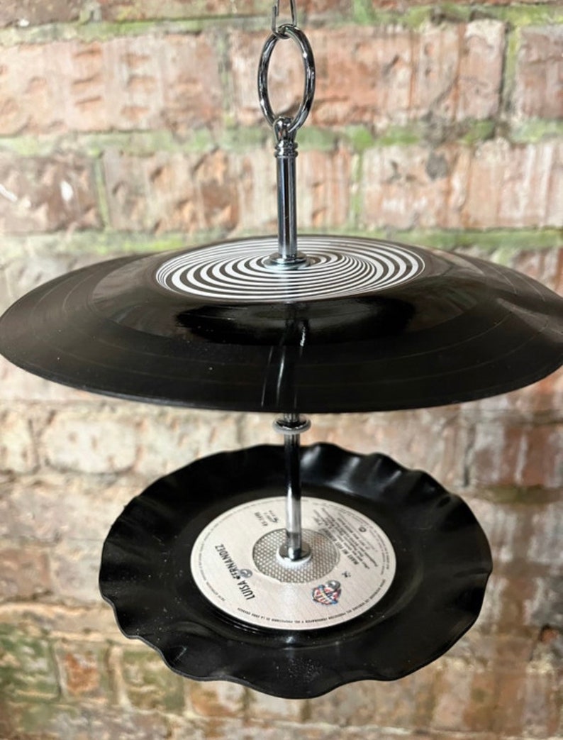 Puede incluir: Un comedero para p&aacute;jaros de dos niveles hecho de discos de vinilo reciclados. El nivel superior presenta un disco negro con un dise&ntilde;o en espiral blanco, mientras que el nivel inferior tiene una etiqueta con texto. Suspendido por una cadena plateada.