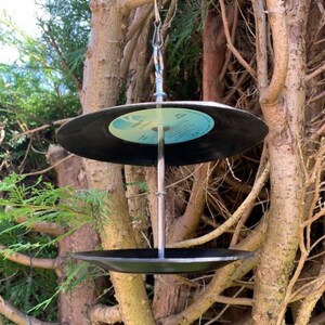 Puede incluir: Un comedero para p&aacute;jaros &uacute;nico hecho de discos de vinilo reciclados. Los discos negros est&aacute;n suspendidos por una cadena y una varilla de metal, con un segundo disco formando la base. El comedero cuelga entre las ramas de los &aacute;rboles.