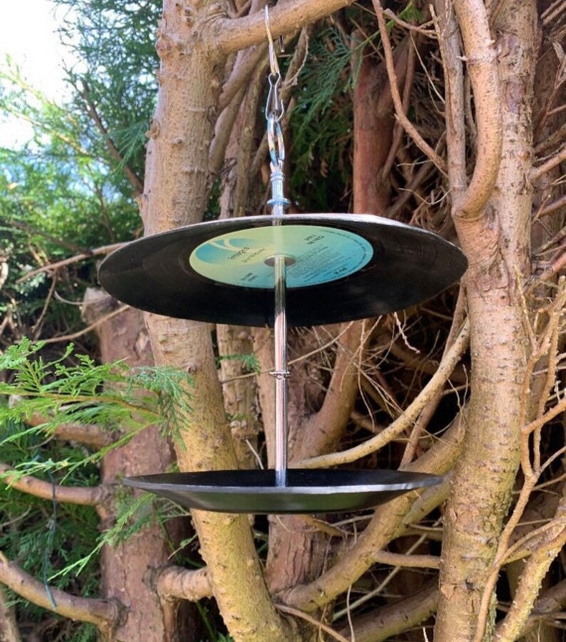 Puede incluir: Un comedero para p&aacute;jaros &uacute;nico hecho de discos de vinilo reciclados. Los discos negros est&aacute;n suspendidos por una cadena y una varilla de metal, con un segundo disco formando la base. El comedero cuelga entre las ramas de los &aacute;rboles.