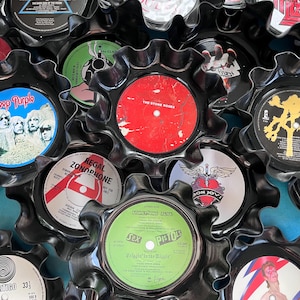 May include: A collection of vintage vinyl record album covers repurposed into coasters. The coasters are black with a scalloped edge and feature a variety of album covers, including Deep Purple, The Stone Roses, Sex Pistols, and Bon Jovi.
