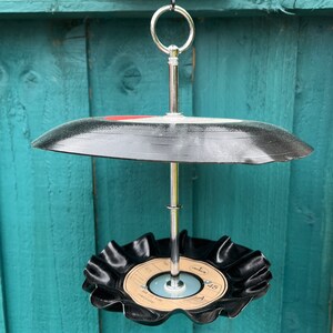 Puede incluir: Un comedero para p&aacute;jaros de dos niveles hecho de discos de vinilo reciclados. Los discos negros est&aacute;n unidos a un soporte de metal plateado con un anillo para colgar. El disco inferior tiene una etiqueta con texto.