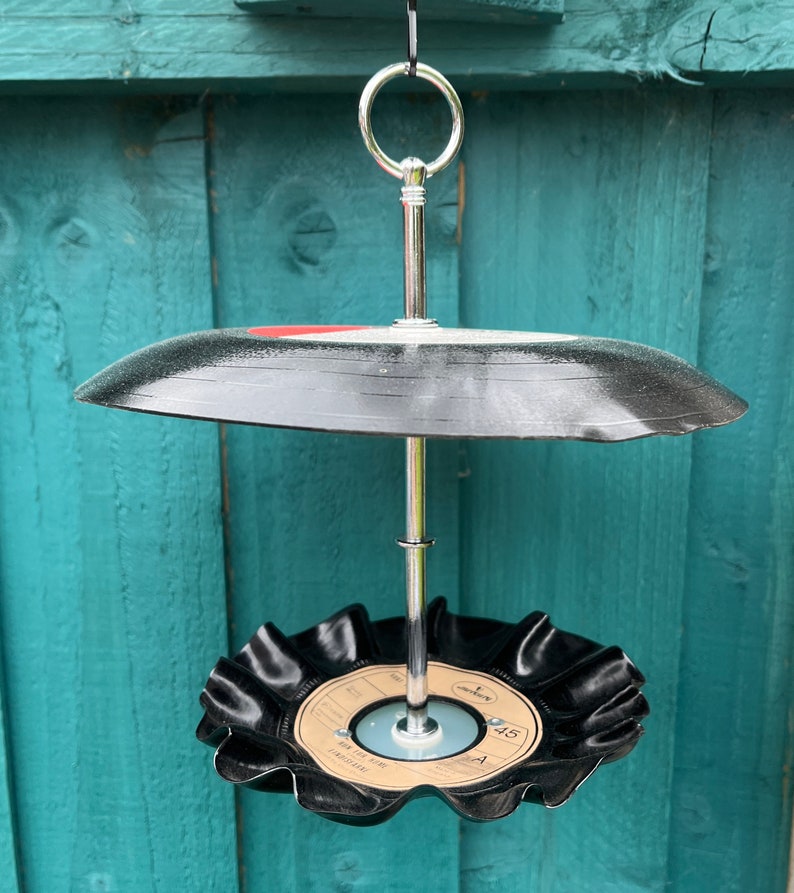 Puede incluir: Un comedero para p&aacute;jaros de dos niveles hecho de discos de vinilo reciclados. Los discos negros est&aacute;n unidos a un soporte de metal plateado con un anillo para colgar. El disco inferior tiene una etiqueta con texto.