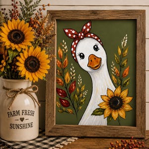 May include: Framed artwork of a goose with a red polka-dot headband, surrounded by autumn leaves and a sunflower. A vase with sunflowers and the text "FARM FRESH SUNSHINE" is on the left. The artwork is in a wooden frame.