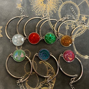 May include: Seven silver bracelets with different colored resin charms. Each charm has a unique design, including a snowflake, a flame, a heart, a flower, a bird, a swirl, and a smiley face.